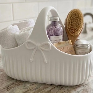 Peut inclure: Un panier en céramique blanc avec un extérieur nervuré et un nœud décoratif sur la poignée. Il contient trois serviettes blanches roulées, une bouteille de sels de bain à la lavande, une brosse de bain en bois, un savon artisanal et un pot de crème.