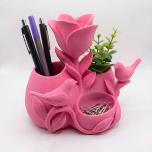 Puede incluir: Un organizador de escritorio de color rosa brillante con una escultura central de rosa, dos figuras de pájaros y tres compartimentos distintos. El organizador contiene varios bolígrafos, una pequeña planta verde y clips metálicos.