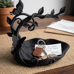 May include: A matte black decorative tray featuring a sculpted tree branch with leaves and two butterflies. The tray holds a set of keys with a brown leather fob, several metallic coins, and a white note with the text for home prints have your Baaly.