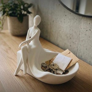 May include: A matte white ceramic sculpture of a seated figure with a smooth, flowing form that extends into a shallow, curved bowl. The bowl contains a small collection of metal keys, several coins, and two small paper envelopes.