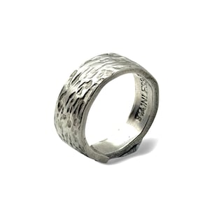 May include: A silver-toned stainless steel ring featuring a wide band with a textured, hammered exterior surface. The interior of the band is smooth and clearly displays the word STAINLESS stamped into the metal surface of the ring.