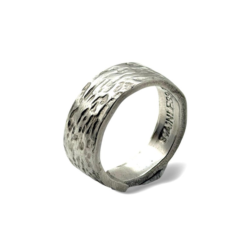 Op de afbeelding: Een zilverkleurige roestvrijstalen ring met een brede band en een getextureerd, gehamerd buitenoppervlak. De binnenkant van de band is glad en toont duidelijk het woord STAINLESS dat in het metalen oppervlak van de ring is gestempeld.