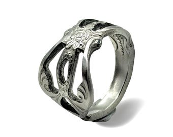 May include: A silver-toned metal ring featuring an openwork design with cutouts and embossed floral patterns. The band has a wide, structured shape with curved edges and a polished finish, showcasing intricate detailing across the surface.
