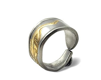 Op de afbeelding: Een zilverkleurige metalen ring met een open, verstelbaar bandontwerp. Het buitenoppervlak heeft een geborstelde textuur en een verdiepte, goudkleurige inleg die een gestileerde vogel of kraanvogel met lange vleugels uitbeeldt.