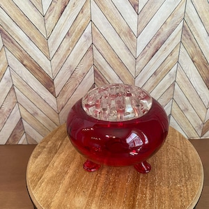 May include: A spherical, ruby red glass flower frog with a clear glass insert. The frog has three small feet and is on a wooden table. The background has a chevron pattern in shades of brown and beige.