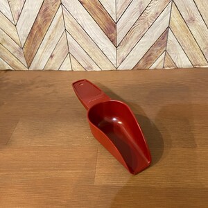 May include: A red plastic scoop with a handle. The scoop is designed for measuring dry ingredients.
