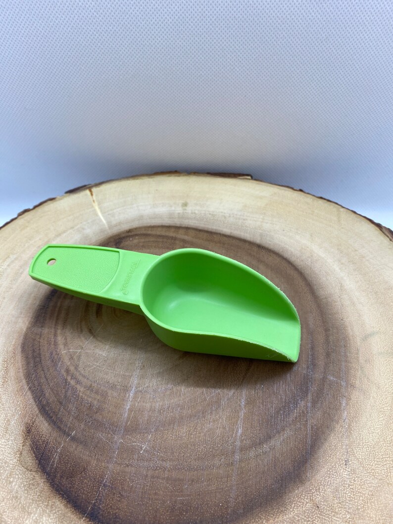 May include: A green plastic scoop with a handle. The scoop is on a wooden surface.