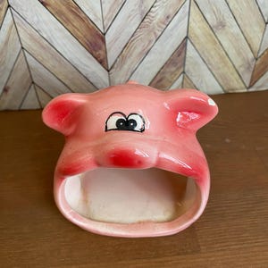 May include: A pink ceramic piggy bank with black eyes and a white spot on the ear. The piggy bank has an opening in the front for coins.