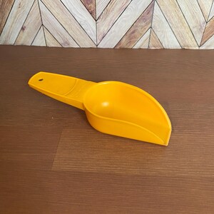 May include: A yellow plastic scoop with a handle. The scoop is designed for measuring dry ingredients.