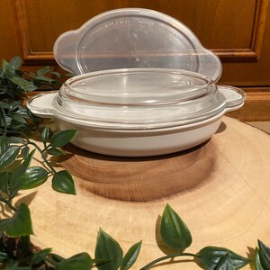 May include: White ceramic baking dish with a clear glass lid. The dish is sitting on a wooden surface.
