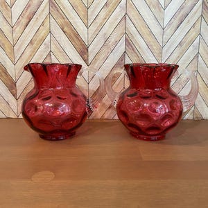 May include: Two ruby red glass jugs with clear, looped handles. The jugs have a ruffled rim and a pattern of raised circles. The jugs are displayed on a wooden surface with a chevron pattern background.