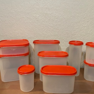 May include: A set of eight clear plastic storage containers with orange lids. The containers are various sizes and shapes, including square and rectangular.