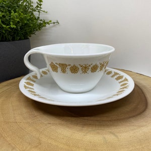 May include: A white teacup and saucer with a gold floral pattern. The cup has a handle and the saucer has a scalloped edge.