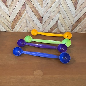 May include: Four melon ball scoops in different colors: yellow, green, purple, and blue. The scoops are lying on a wooden surface.