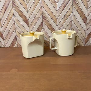 May include: Two off-white, retro-style kitchen canisters with yellow lids. One canister has a handle, the other does not. The canisters have a unique, angular design. The background is a wooden chevron pattern.