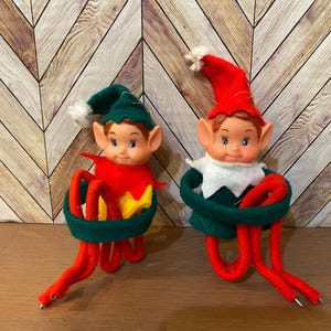 May include: Two vintage elf ornaments. Each elf has a plastic face with blue eyes, red hair, and a pointed hat. One elf wears a red hat and the other wears a green hat. Both have a green and red outfit with red legs.