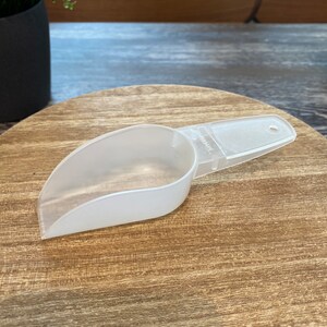 May include: A white plastic scoop with a handle. The scoop is on a wooden surface.