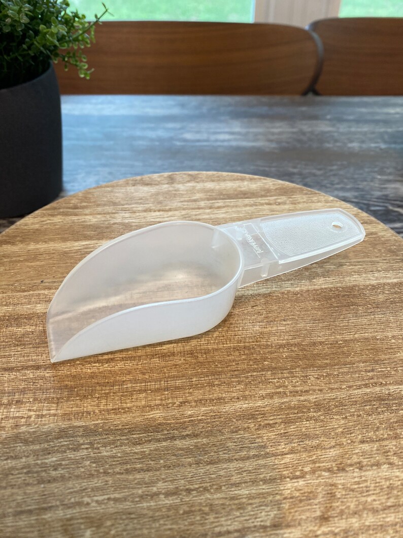 May include: A white plastic scoop with a handle. The scoop is on a wooden surface.