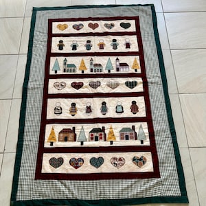 May include: A rectangular quilt featuring horizontal rows of appliqued hearts, stylised figures, and houses. The design includes a cream background with deep red borders and a green and white chequered outer frame with a solid dark green edge.