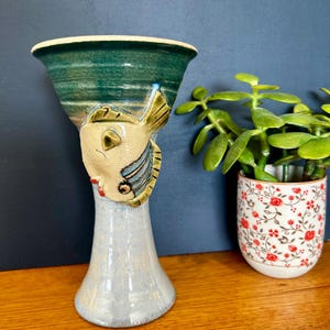 May include: A ceramic goblet featuring a flared green rim and a light blue base. A sculpted, beige-coloured fish with blue-striped fins and red lips is attached to the side of the stem, positioned between the bowl and the base of the vessel.