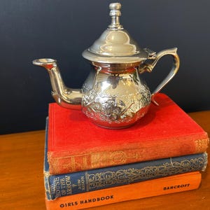 May include: A polished silver-toned metal teapot with an ornate embossed floral pattern, a curved spout, a hinged lid with a finial, and a handle. The teapot rests on a stack of three vintage hardcover books with red, blue, and orange spines.