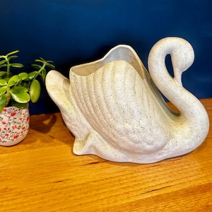 May include: A ceramic swan-shaped planter featuring a cream-coloured base with a speckled brown texture. The swan has a curved neck and moulded wing details on its body, with an open top section designed to hold small plants or decorative items.