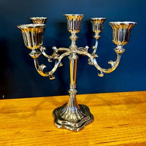 May include: A polished silver-tone metal candelabra featuring five candle holders arranged in a tiered design. The central holder is elevated above four curved arms, each ending in a fluted cup. The base is wide, scalloped, and reflective.