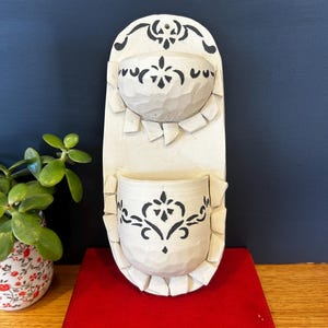 May include: A vertical wall-mounted ceramic planter featuring two stacked, semi-circular pockets. The off-white surface is decorated with black stencilled floral patterns and has a textured, hand-carved appearance with small decorative clay tabs.