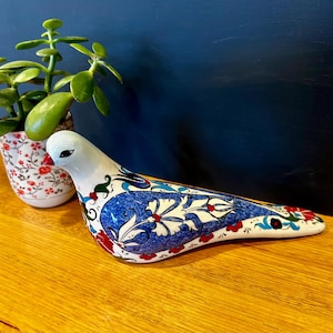 May include: A ceramic bird figurine featuring a white base with intricate blue, red, and green floral patterns. The bird has a smooth, glossy finish, a pointed red beak, and detailed painted motifs covering its body, wings, and tail feathers.