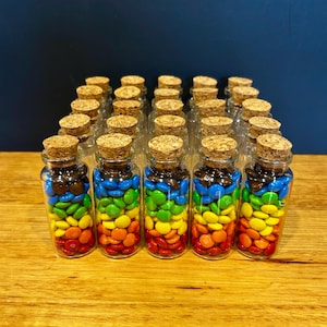 May include: A collection of twenty-five small, clear glass bottles arranged in a square grid. Each bottle is filled with layers of colourful, round, candy-coated chocolates in red, orange, yellow, green, blue, and brown, topped with a cork stopper.