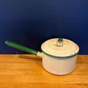 May include: A cream-coloured enamel saucepan with a long green handle and a matching lid. The pot features a green rim around the top edge and a green knob on the centre of the lid. The exterior surface shows signs of wear with minor chips.