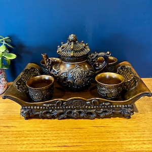 May include: A bronze-coloured tea set featuring a teapot with a detailed lid and handle, accompanied by four matching cups, all arranged on a rectangular tray with ornate, raised edges and intricate relief carvings throughout the set.