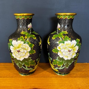 May include: A pair of black cloisonné vases featuring a repeating wire pattern, large white and yellow peony flowers with green leaves, and a gold-toned rim. The vases have a narrow neck that flares at the top and a rounded body with a floral design.
