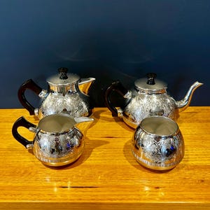 May include: A four-piece silver-plated tea set comprising two teapots, a creamer, and a sugar bowl. Each item features a polished metallic finish with detailed floral engravings, dark brown handles, and dark brown knobs on the teapot lids.