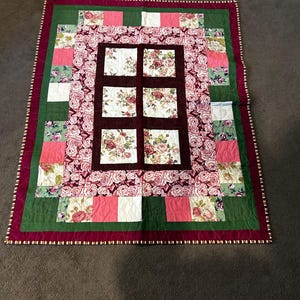 May include: A rectangular patchwork quilt featuring a central grid of six floral squares framed by deep burgundy borders. The design includes alternating blocks of pink, cream, and green fabric with rose patterns, finished with a dark red outer edge.