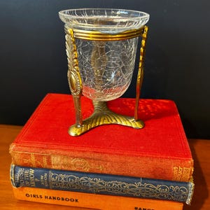 May include: A clear crackle glass vessel held within a gold-toned metal stand featuring three ornate legs. The stand has a circular rim and a base with leaf-like detailing. The vessel is positioned atop a stack of three hardcover books.