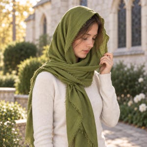 May include: An olive green scarf with a frayed edge finish draped over the head and shoulders. The scarf is made of a lightweight, textured fabric and is worn over a plain, long-sleeved, cream-colored knit sweater with a ribbed texture.