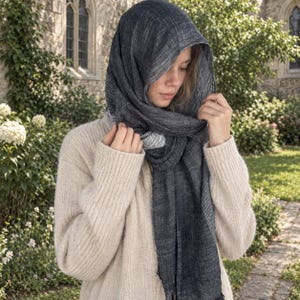 May include: A dark charcoal gray scarf with a subtle white pinstripe pattern draped over the head and shoulders, paired with a beige long-sleeved knit sweater featuring a ribbed texture on the sleeves and a solid knit body construction.