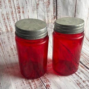 May include: Two cylindrical glass jars with a vibrant red translucent finish. Each jar features a circular, threaded metal lid with a weathered, matte gray texture. The jars are positioned side by side, showcasing their smooth, uniform glass walls.
