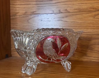 Puede incluir: Un cuenco de vidrio prensado transparente con un patrón de diamantes y tres pies ornamentados. El centro del cuenco muestra un medallón circular rojo con un relieve de un pájaro posado en una rama junto a dos espigas de trigo.