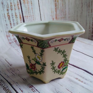 May include: A hexagonal ceramic planter with a cream-colored base, featuring hand-painted floral motifs in red, white, and green. The rim is decorated with a green lattice pattern and small red flowers, while the body displays trailing vines.