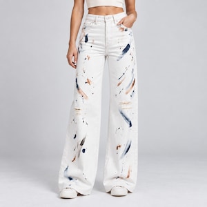 May include: High-waisted white denim jeans featuring a wide-leg silhouette. The fabric is decorated with abstract paint splatters and long, sweeping brushstrokes in shades of navy blue, light blue, and tan, creating a textured artistic effect.