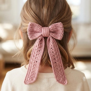 May include: A dusty rose pink crochet hair bow featuring a central knot and two long, dangling ribbon-like tails. The accessory is crafted with a textured yarn stitch pattern, creating a soft, dimensional appearance for securing hair in place.