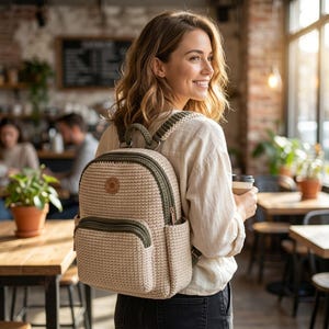 Puede incluir: Una mochila de ganchillo en color beige con ribetes verde oliva, que cuenta con un bolsillo frontal con cremallera, un asa superior y correas ajustables. La bolsa tiene un parche circular con un logotipo marrón en la parte delantera.