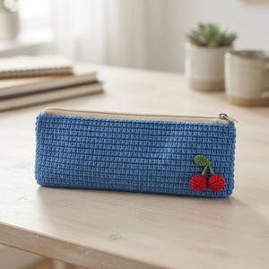 May include: A rectangular blue crochet pencil case featuring a small, attached red cherry motif with green stems and leaves. The case is secured with a beige zipper closure along the top edge, providing a structured storage solution for stationery items.