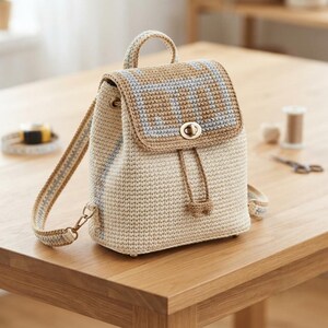 May include: A crochet backpack in cream and tan with a light blue geometric pattern on the flap. The bag features a gold-tone metal turn-lock closure, a top handle, adjustable shoulder straps, and a drawstring cord with knotted ends.