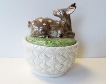 May include: A ceramic decorative jar with a deer figurine on top. The deer is brown with white spots and is lying on a green base. The jar is white with a woven basket design.