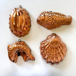 May include: A set of four copper-colored metal molds featuring distinct shapes including an oval fluted design, a fish, a curved dolphin or sea creature, and a large scallop shell. Each mold includes a small metal loop at the top for hanging.