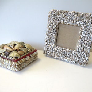 May include: A decorative box and a picture frame, both adorned with seashells. The box has a red border and a lid covered in various shells. The frame is square, with a neutral-colored backing, and is completely covered in small, spiral shells.
