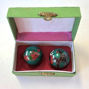 May include: Two spherical metal balls with green enamel and intricate black and red dragon designs, housed in a rectangular green box with a gold-toned metal latch and a red velvet-lined interior. The box lid is open, revealing the two spheres.